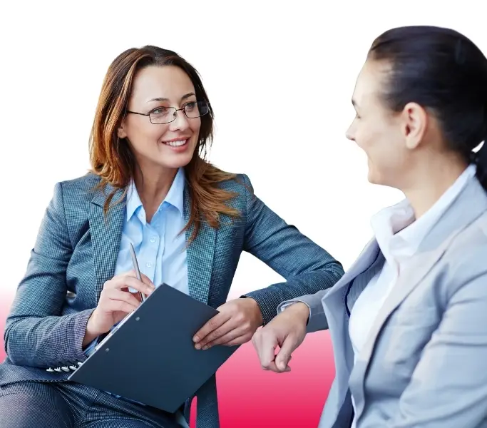 Zwei Frauen in Anzügen vor hellem Hintergrund, die sich ansehen, wobei eine davon eine Brille trägt und aufmerksam zuhört.